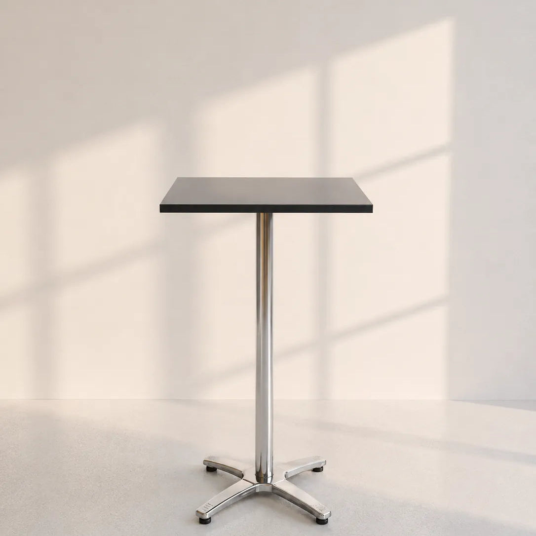 Image of A modern square bar table with a black top and a polished chrome pedestal base, set against a minimalist off-white wall with soft window shadow patterns.