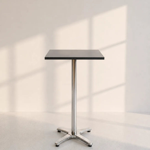A modern square bar table with a black top and a polished chrome pedestal base, set against a minimalist off-white wall with soft window shadow patterns.