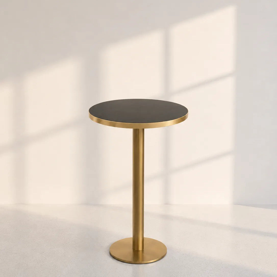 A modern round side table with a dark charcoal tabletop and a brushed gold metal pedestal base, set against a minimalist off-white wall with soft window shadow patterns.