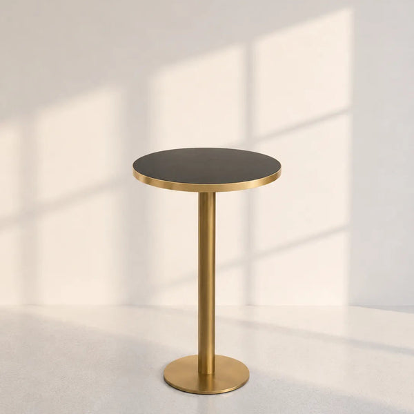 A modern round side table with a dark charcoal tabletop and a brushed gold metal pedestal base, set against a minimalist off-white wall with soft window shadow patterns.
