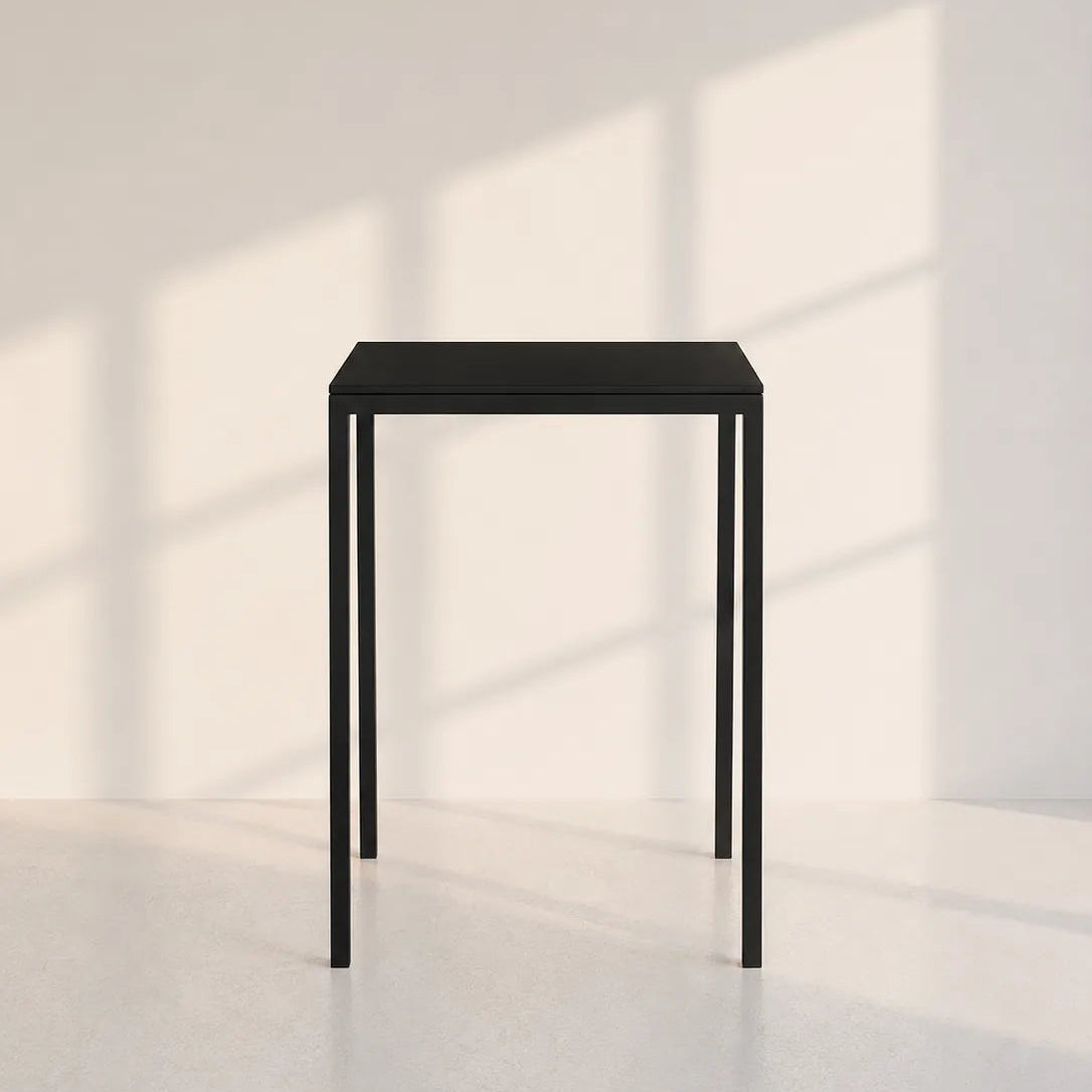 A minimalist black metal side table with a square top and four slim legs, positioned in a bright, modern room with soft window shadow patterns on the wall and floor.