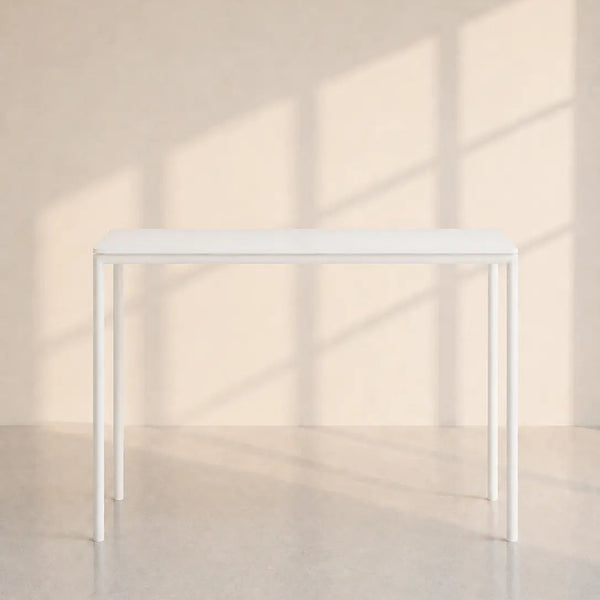 A minimalist white rectangular table with slim legs, positioned against a warm beige wall with soft window shadow patterns.