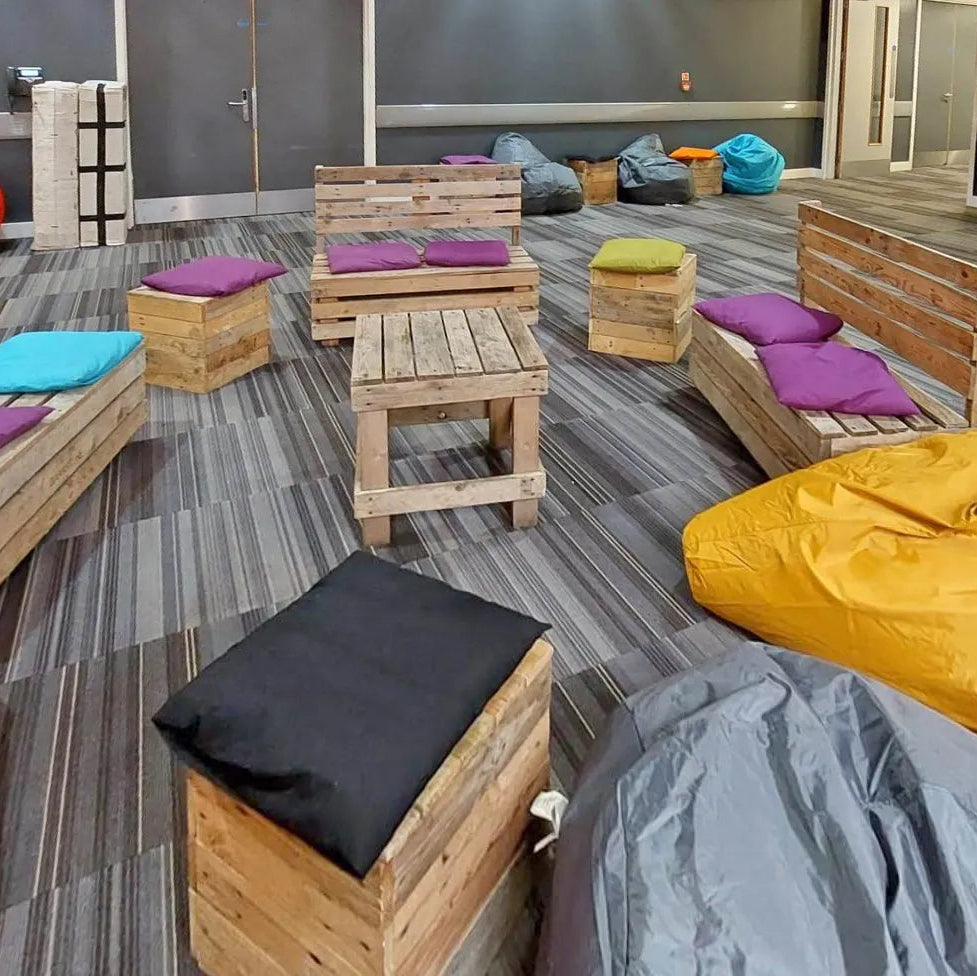 A lounge area with wooden pallet-style seating and bean bags in orange, yellow, gray, black, purple, and turquoise on a gray striped floor.