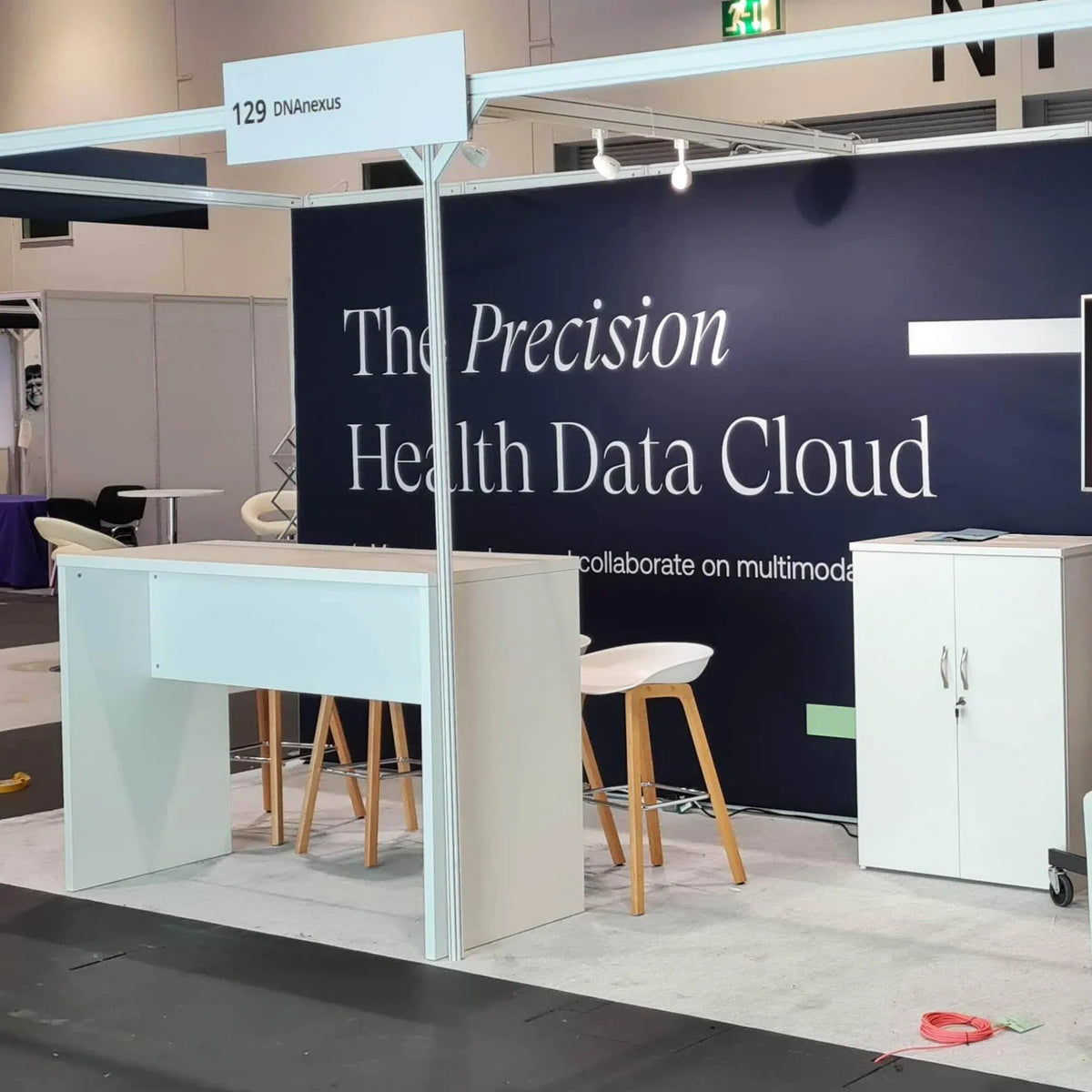 A booth display at a trade show featuring a dark navy blue wall with the text 'The Precision Health Data Cloud' in white, white tables and stools, a mounted black screen, and cream-colored curved chairs under a white overhead frame.