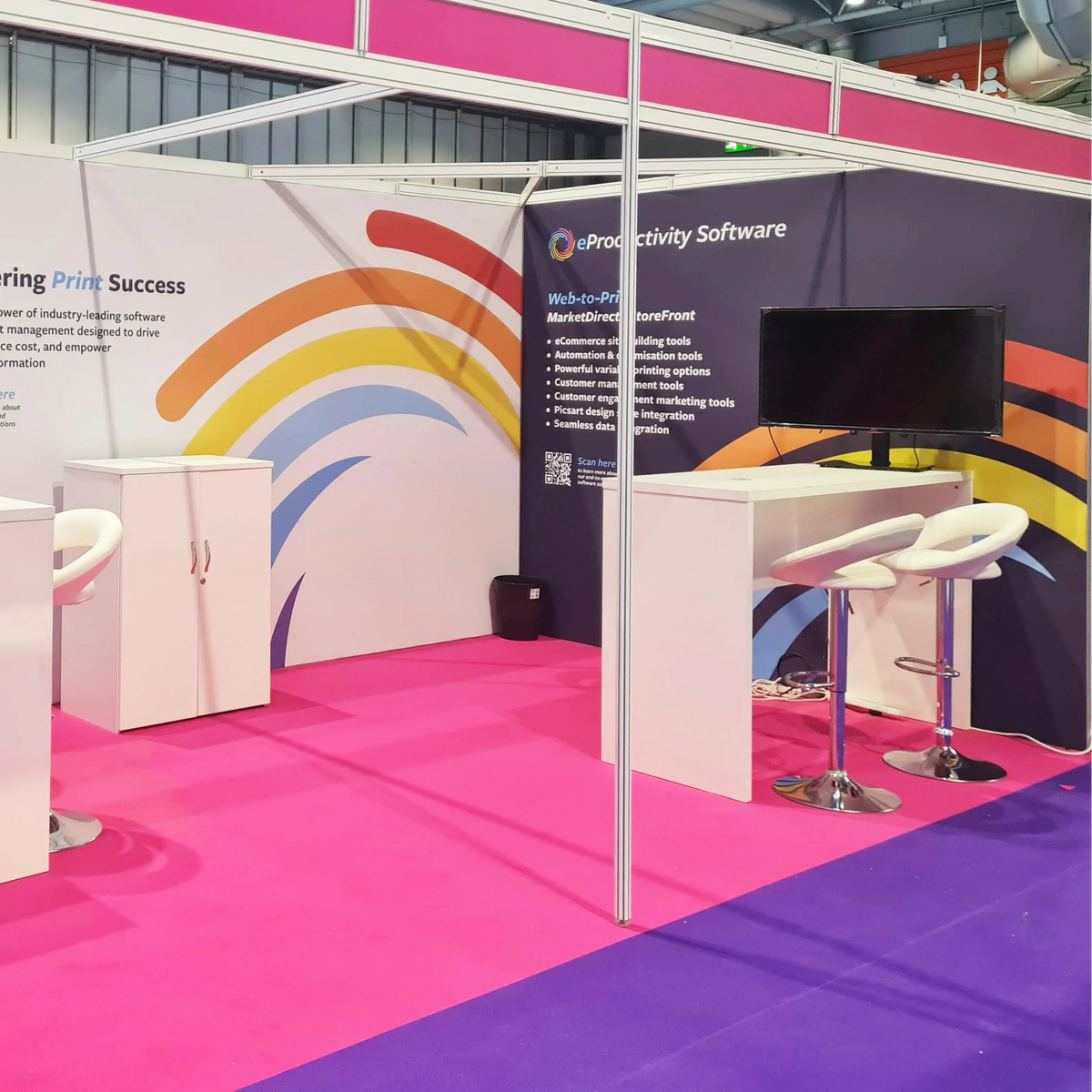 A booth display for 'ubiquity Software' at a trade show, featuring white desks with white bar stools, computer monitors, and a colorful rainbow graphic on a white and dark blue backdrop.