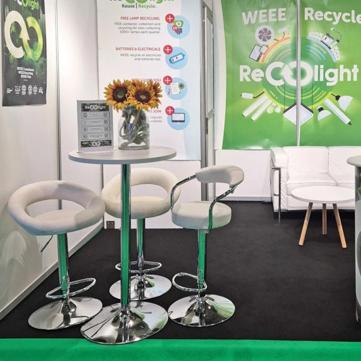 Image of A booth display for ReCOlight featuring a green and white branded banner, a curved reception desk with recycling graphics, a small round table with three white swivel bar stools, and a white sofa in the background.