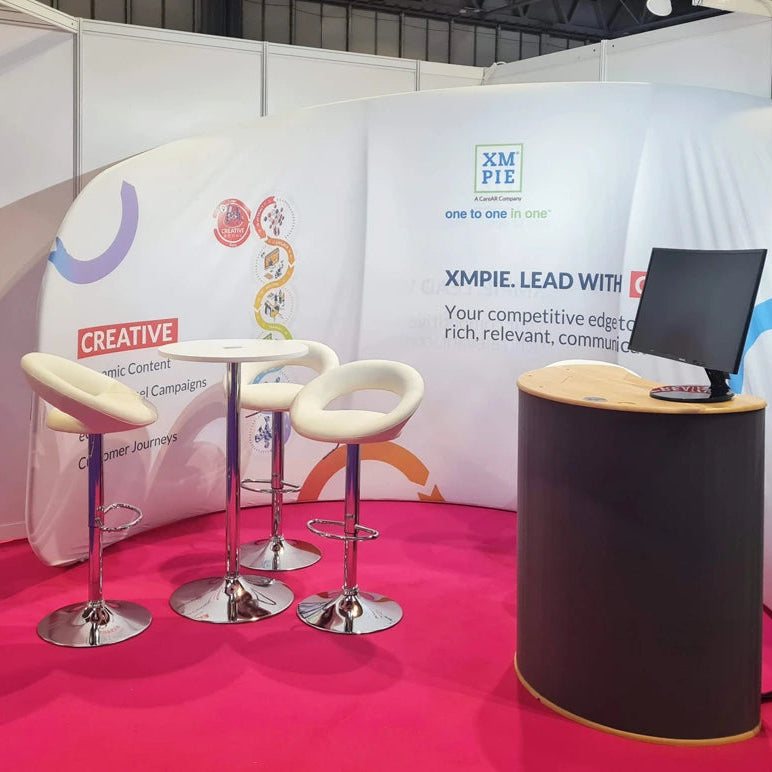 Image of A promotional booth setup with a curved white backdrop featuring colorful graphics and text, including XM PIZ and XMPIE LEAD WITH. The setup includes a small round table with three cream-colored bar stools, a black computer monitor on a cylindrical black-and-wood counter, and a bright pink floor covering.
