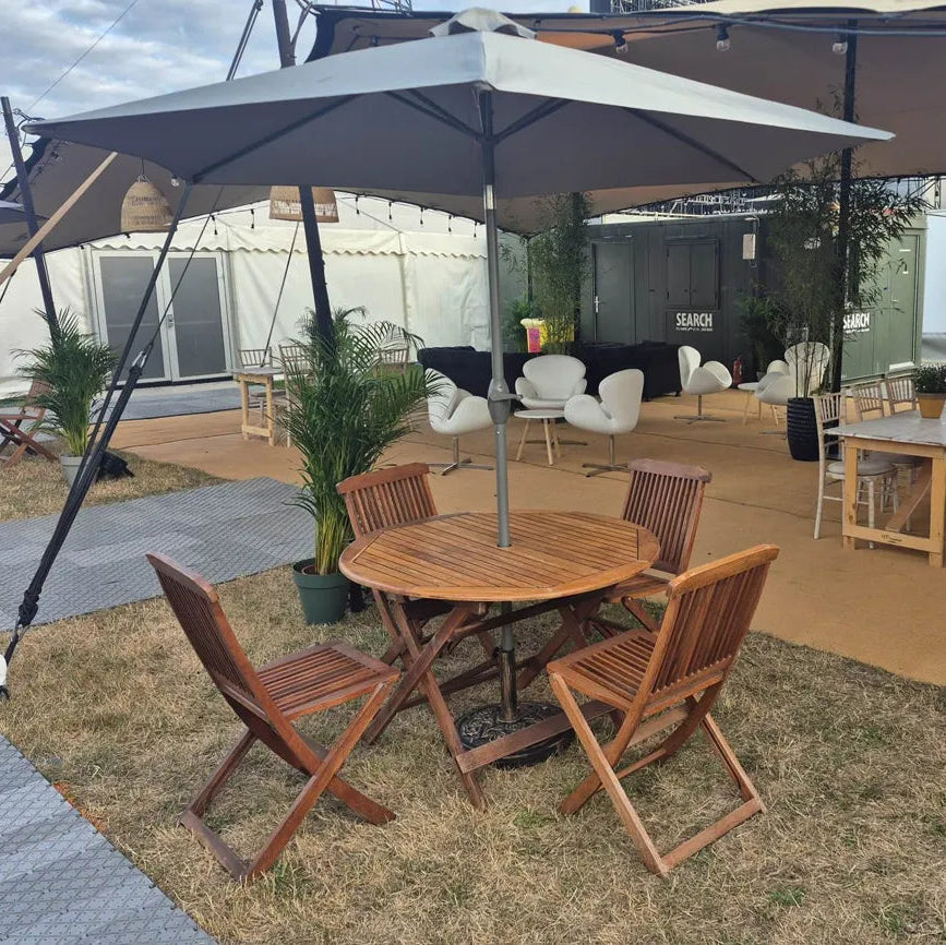 Image of A round wooden folding table with four matching wooden folding chairs under a large gray patio umbrella, set on a grassy outdoor event area with tents and lounge furniture in the background.