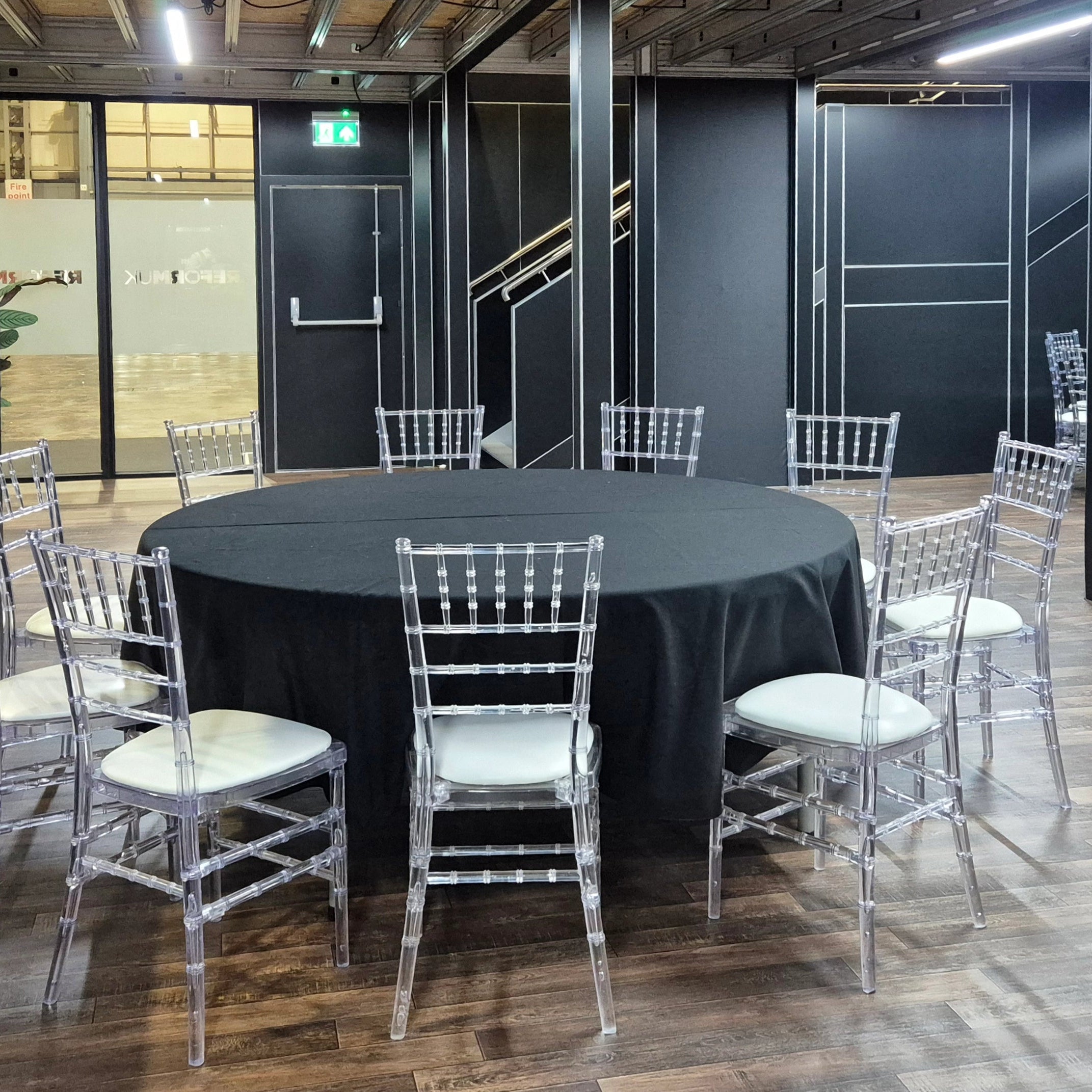 Image of A round black tablecloth-covered dining table surrounded by clear acrylic Chiavari chairs with white upholstered seats in a modern event space with dark walls and wood flooring.