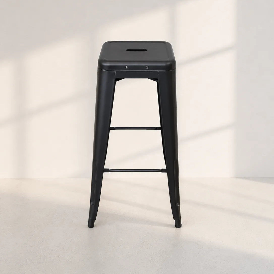 A black metal bar stool with a square seat, backrest cutout, and two lower support bars, photographed in a minimalist studio setting with soft window-box shadows on a light wall and floor.