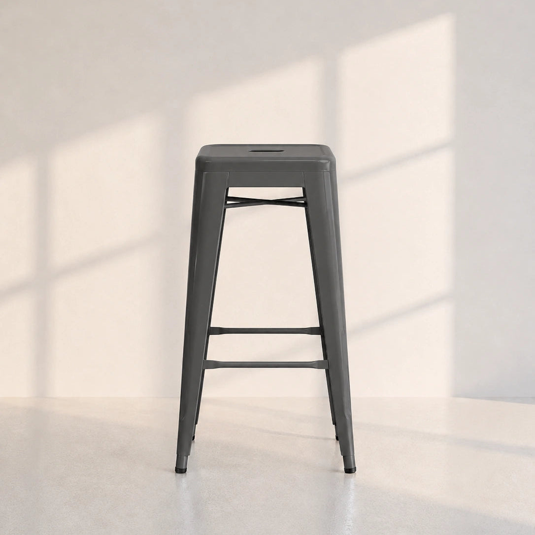 A dark gray metal bar stool with a square seat and four legs, positioned in a minimalist room with soft window shadow patterns on the wall and floor.