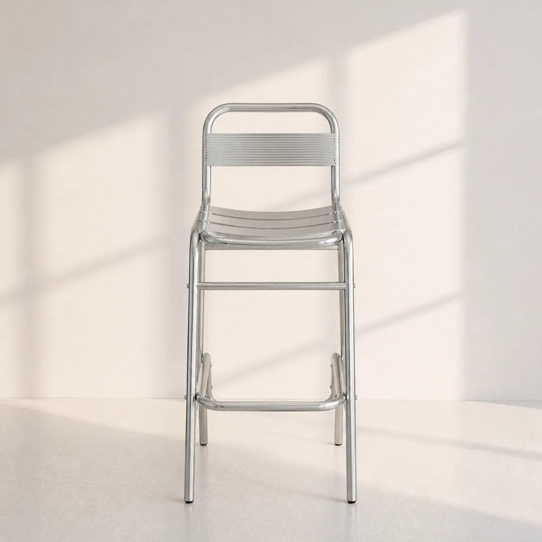 Image of A modern metal bar stool with a silver aluminum frame, featuring a ribbed seat and backrest, set against a minimalist off-white wall with soft window shadow patterns.
