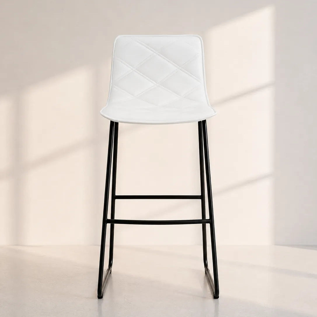 Image of A modern bar stool with a white quilted faux-leather seat and slim black metal legs, set against a minimalist beige wall with soft window shadow patterns.