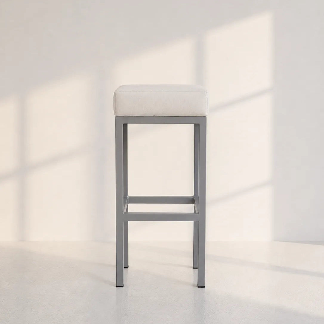 A modern bar stool with a light beige upholstered cushion and a gray metal frame, set against a minimalist white wall with soft window shadow patterns.