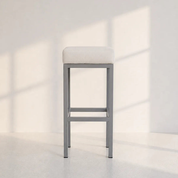 A modern bar stool with a light beige upholstered cushion and a gray metal frame, set against a minimalist white wall with soft window shadow patterns.