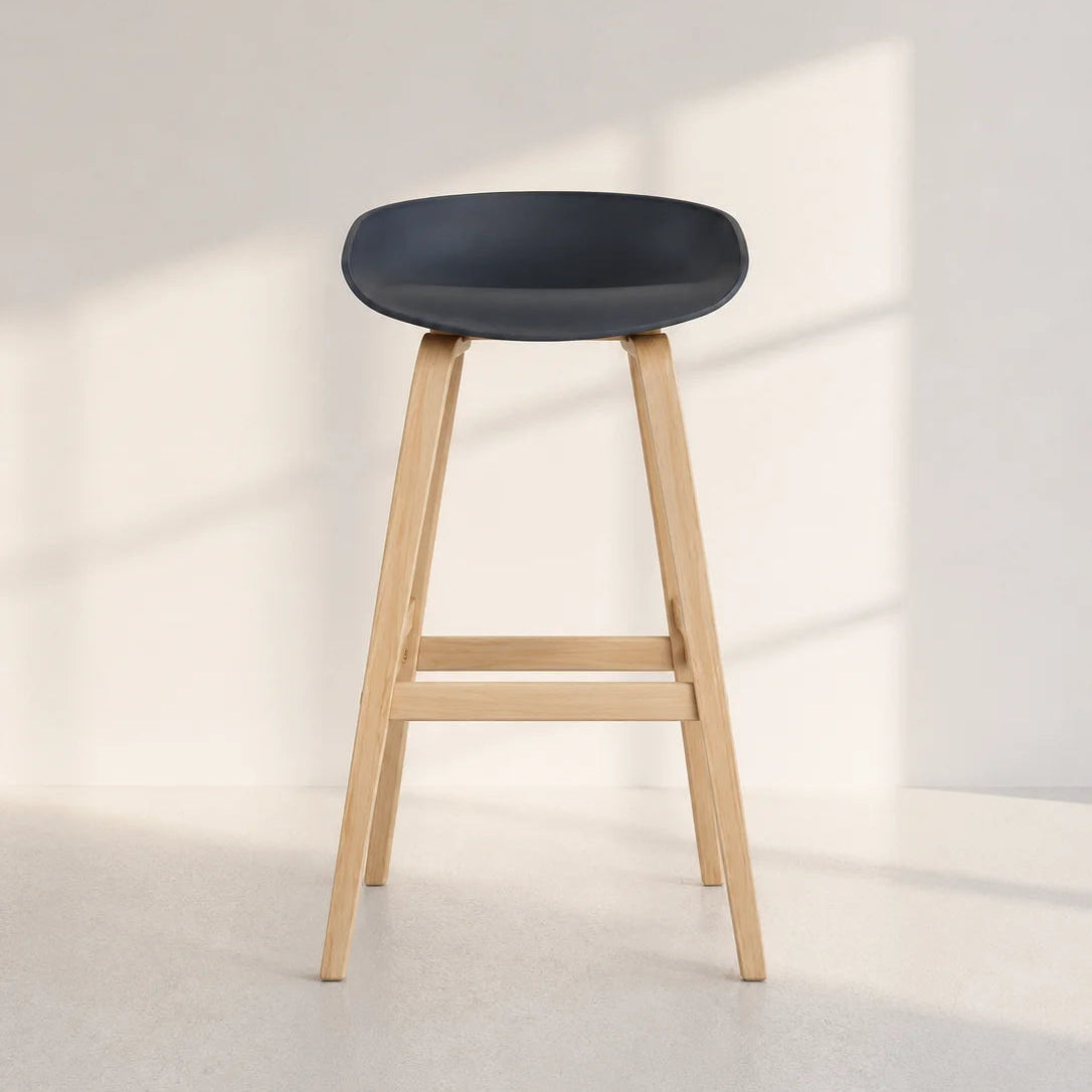Image of A modern bar stool with a dark charcoal gray molded seat and light natural wood legs, set against a minimalist off-white wall with soft window shadows.