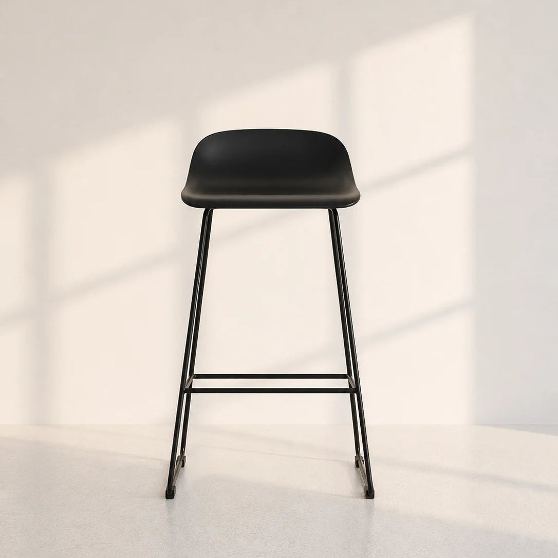 A modern black bar stool with a curved seat and slim metal legs, positioned against a minimalist off-white wall with soft window shadow patterns.