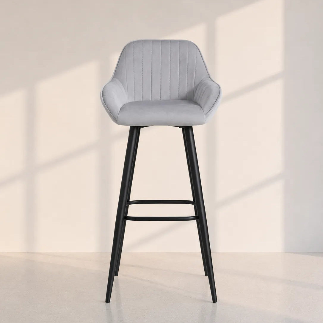 Image of A modern bar stool with a light gray velvet upholstered seat and a black metal frame, set against a minimalist beige wall with soft window shadow patterns.