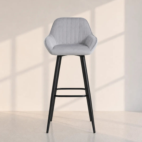A modern bar stool with a light gray velvet upholstered seat and a black metal frame, set against a minimalist beige wall with soft window shadow patterns.