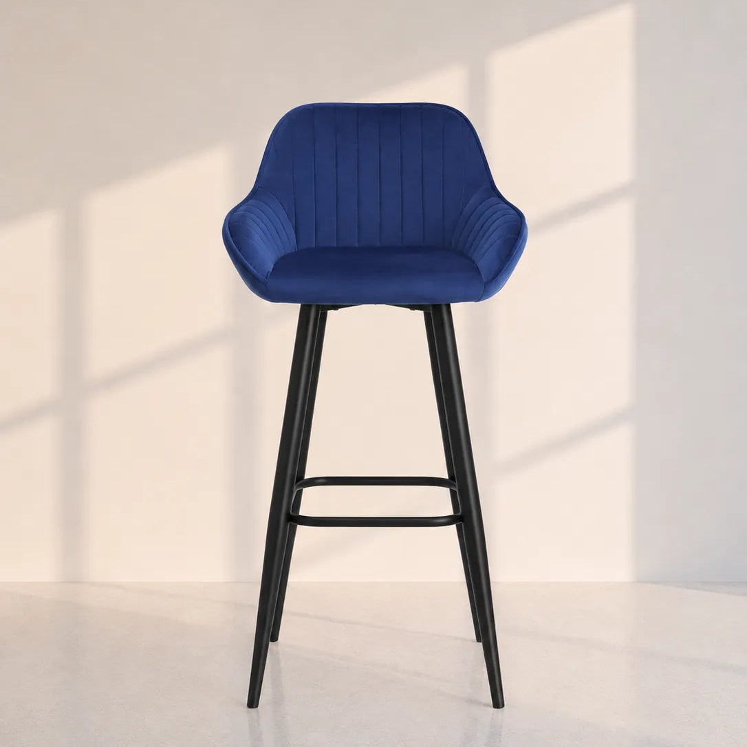 Image of A modern bar stool with a deep blue ribbed upholstered seat and backrest, supported by slim black metal legs, set against a minimalist off-white wall with soft window shadow patterns.