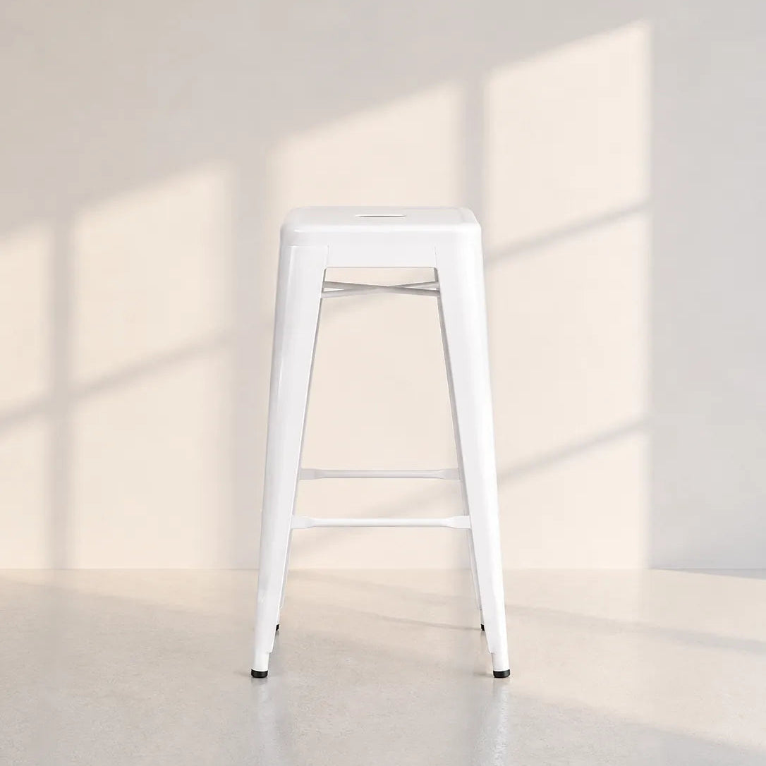 A white metal high stool with a square seat and two front support bars, photographed in a minimalist room with soft window-box shadows on a light wall and floor.