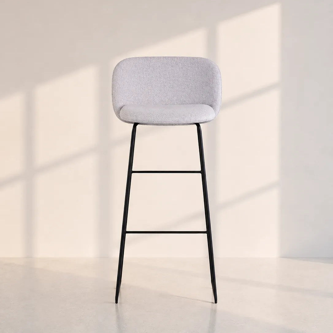Image of A modern bar stool with a light gray textured fabric seat and a slim black metal frame, set against a minimalist beige wall with soft window shadow patterns.