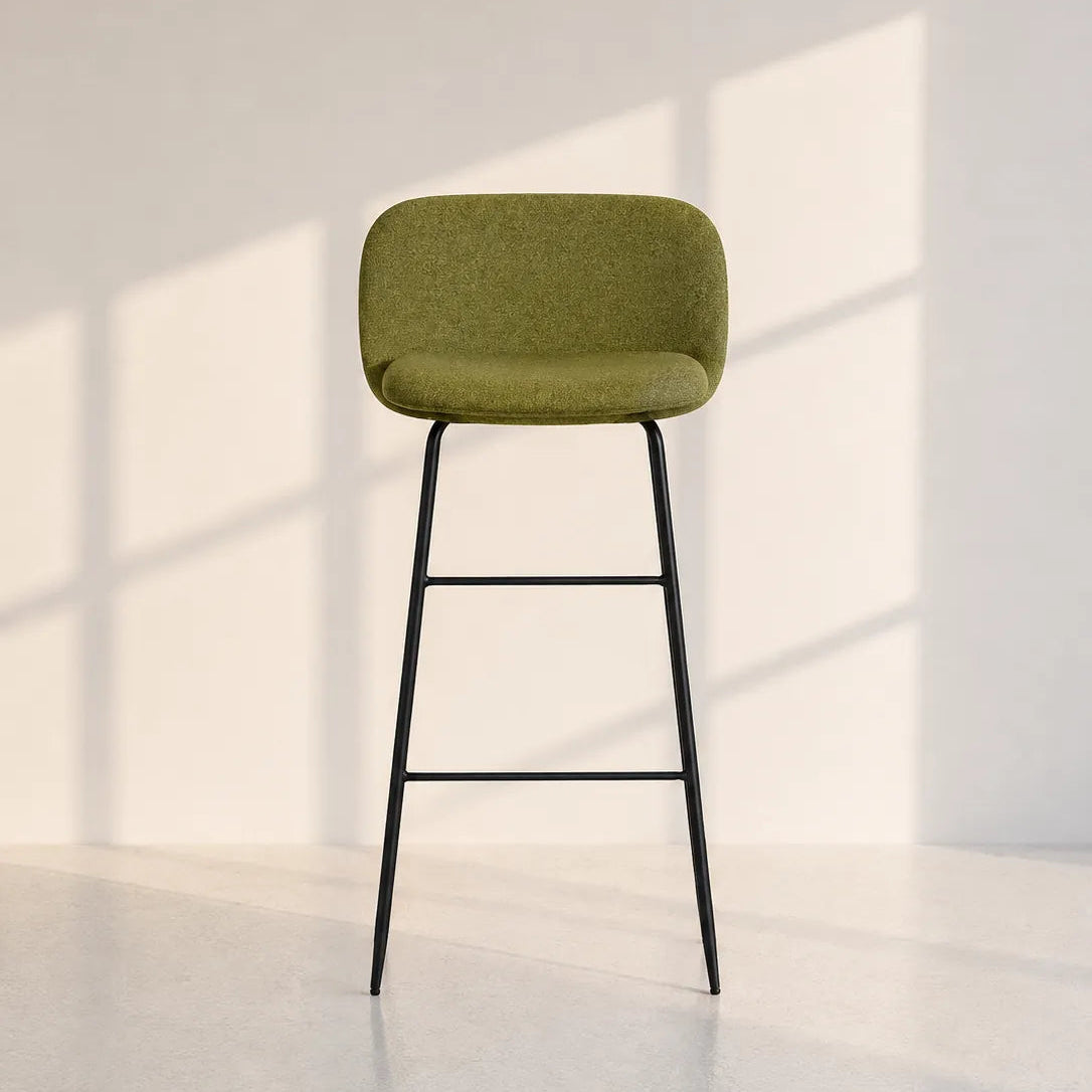 A modern bar stool with an olive green upholstered seat and backrest on a slim black metal frame, set against a minimalist white wall with soft window shadow patterns.
