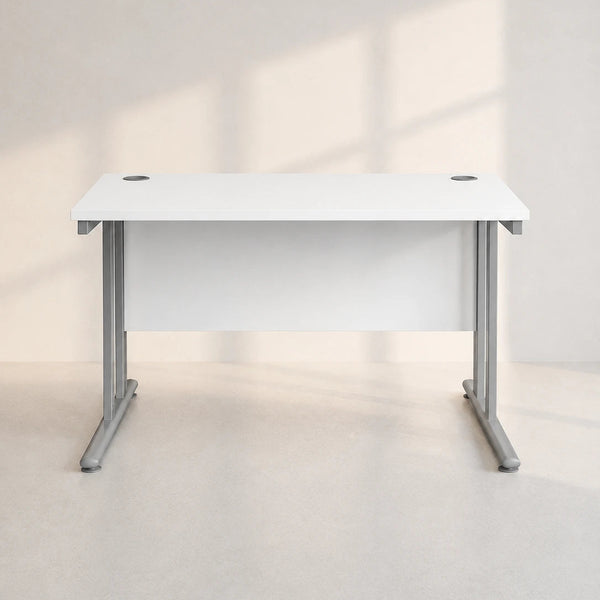 A modern white desk with a white top and gray metal legs, shown in a minimalist room with soft window shadows.