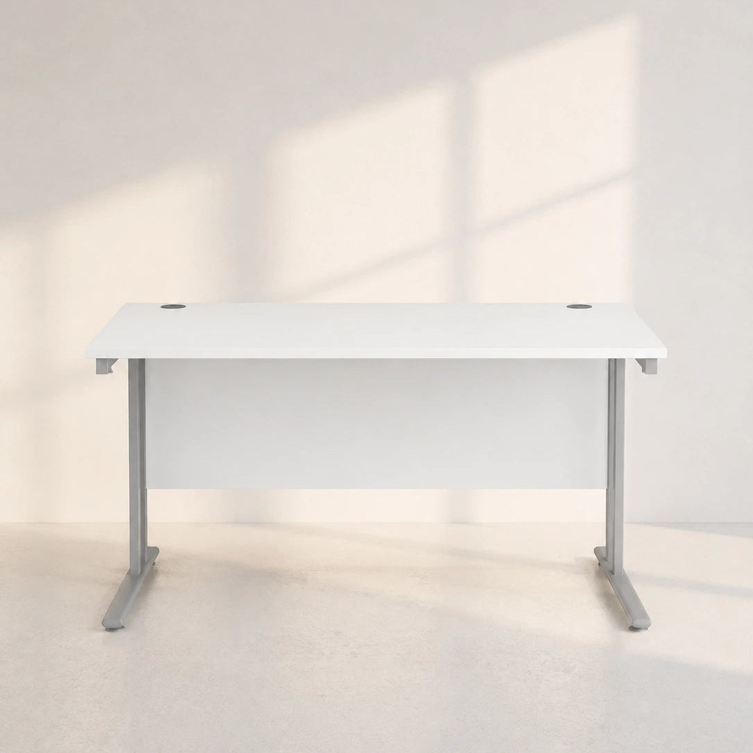 A modern white desk with a white desktop and gray metal legs, positioned against a minimalist off-white wall with soft window shadows.