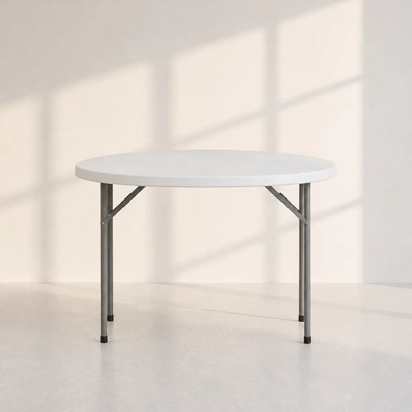 A round folding table with a white top and gray metal legs, set against a minimalist off-white wall with soft window shadow patterns.