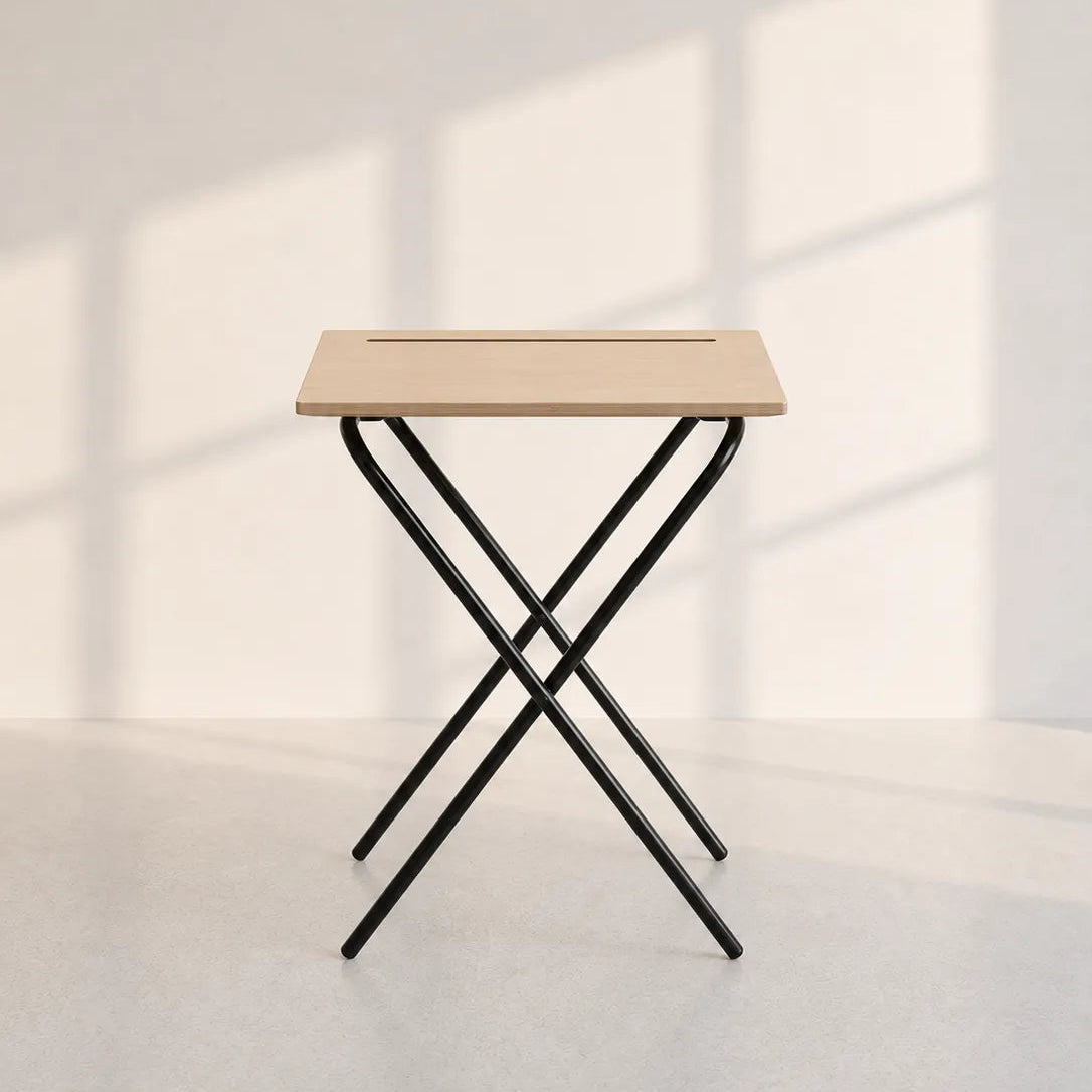 A small folding table with a light natural wood top and black metal X-frame legs, photographed in a minimalist room with soft window shadows on a white wall.