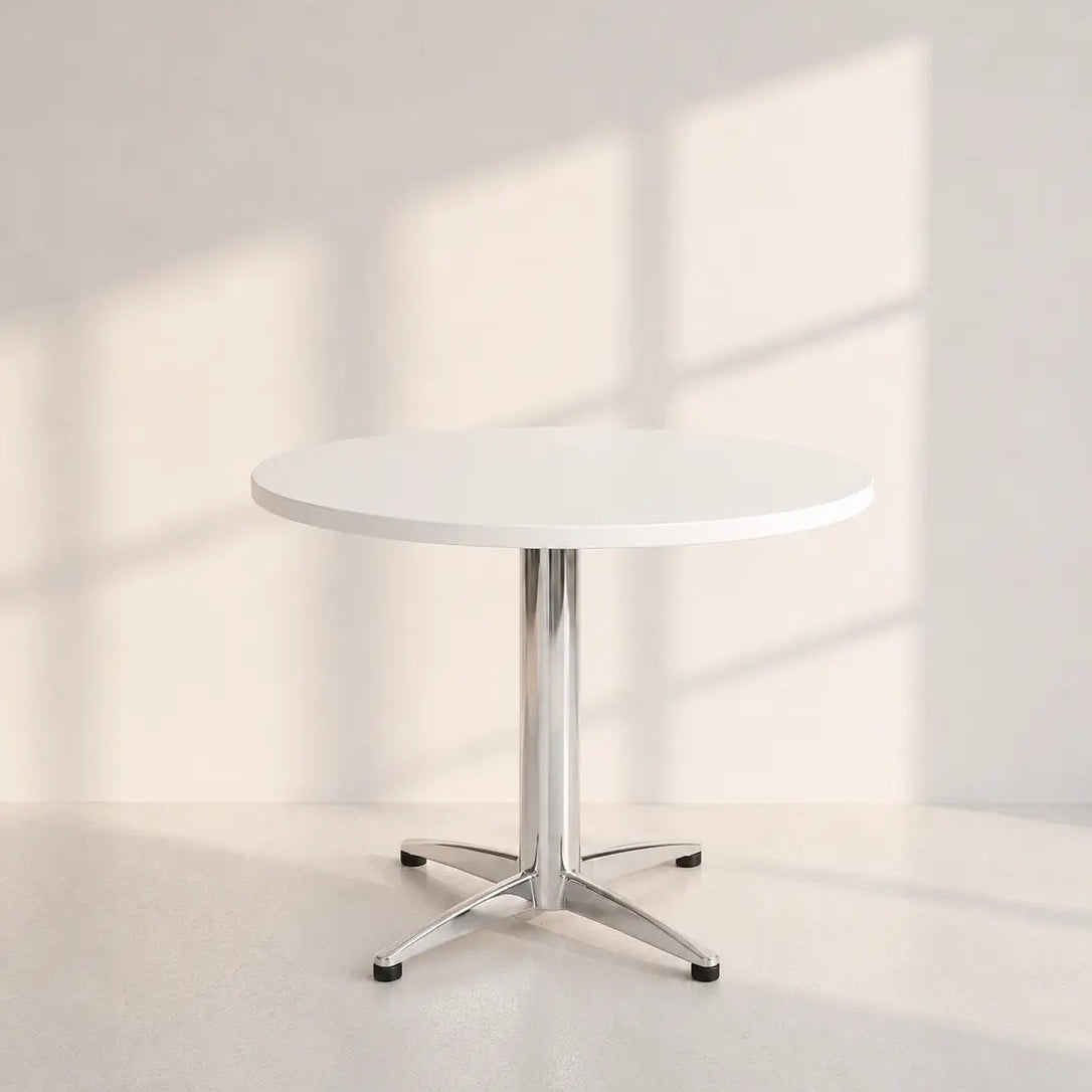 Image of A minimalist round white table with a chrome pedestal base and star-style legs, set against a warm off-white wall with soft window shadow patterns.
