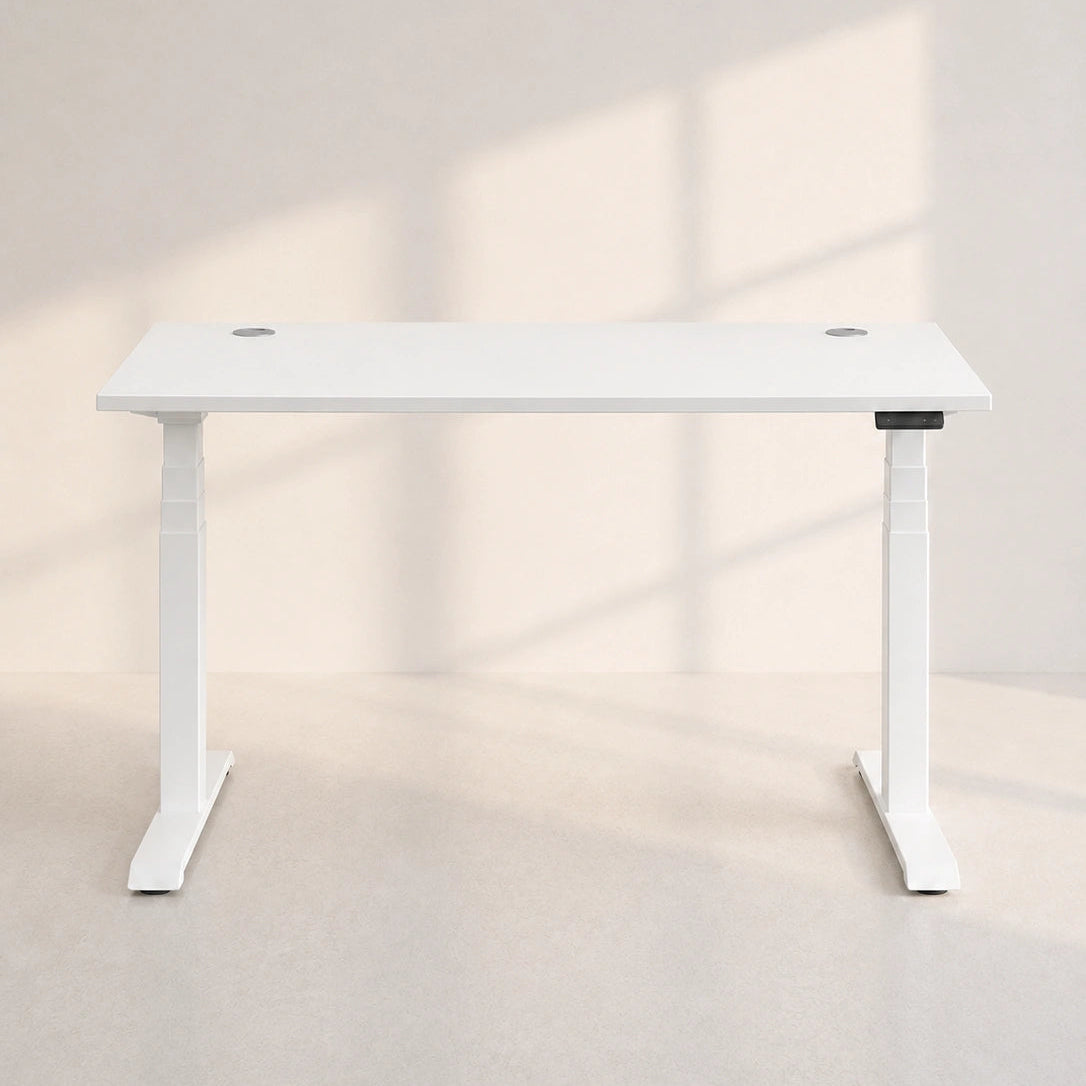 Image of A minimalist white standing desk with a rectangular top and T-style legs, shown in a clean studio setting with soft window-shadow lighting on a light beige wall and floor.