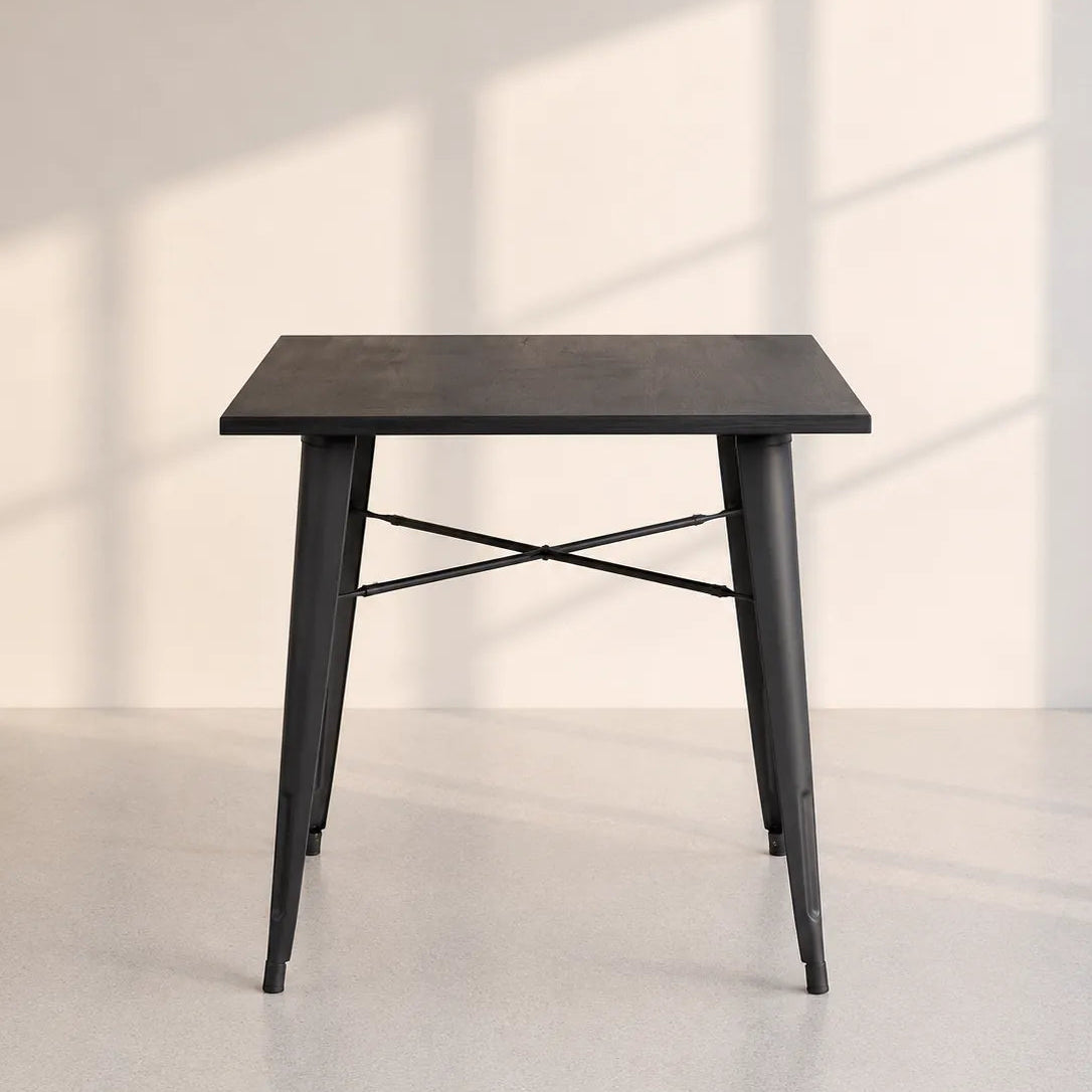 A square black table with tapered metal legs and an X-shaped crossbar base, photographed in a minimalist room with soft window shadow patterns on the wall.
