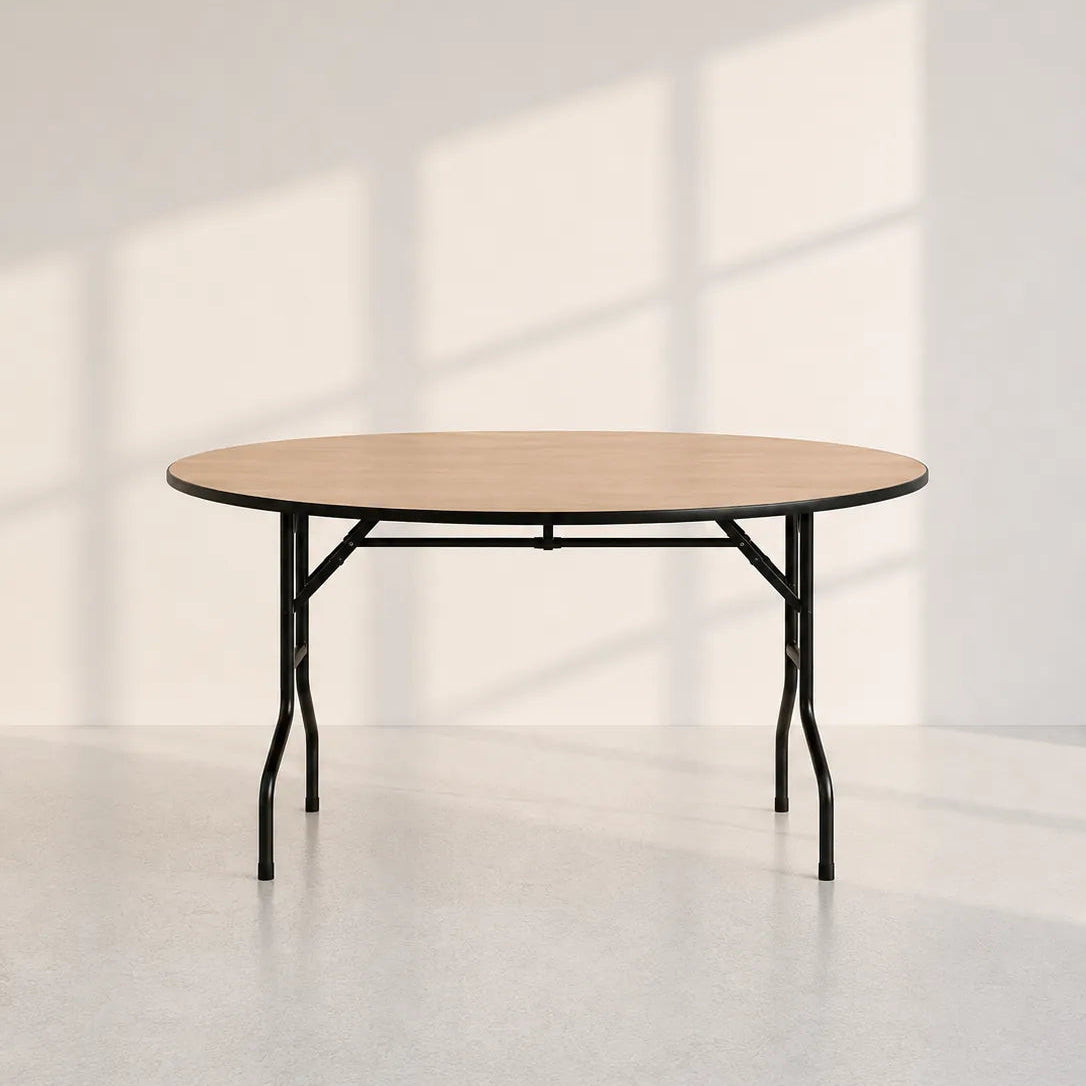 A round folding table with a light natural wood top and black metal legs, set against a minimalist off-white wall with soft window shadow patterns.