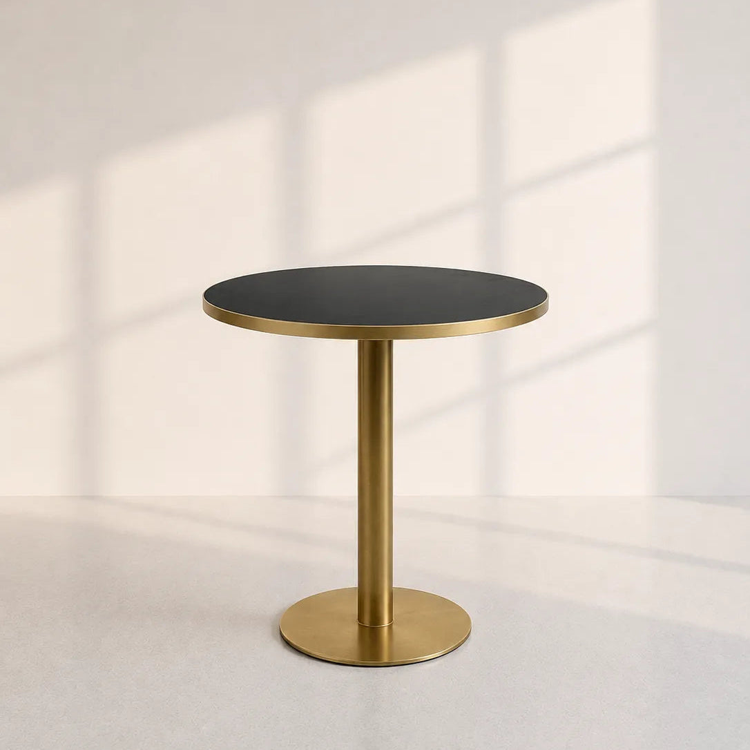 Image of A modern round side table with a matte black top and a brushed gold pedestal base, set against a light gray wall with soft window shadow patterns.