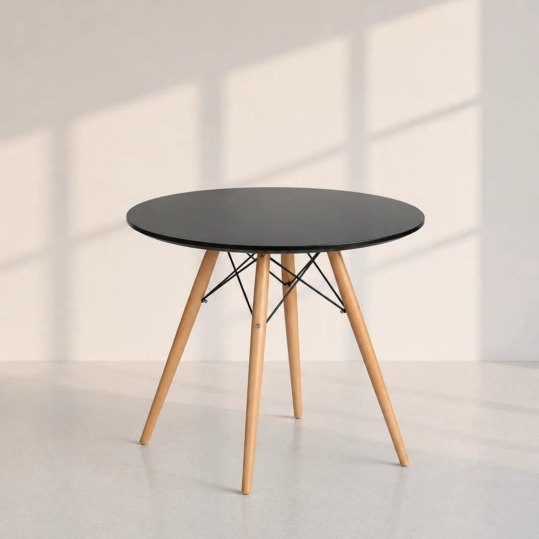 Image of A modern round side table with a black top and light natural wood legs, supported by thin black wire crisscrossing at the center, set against a minimalist off-white wall with soft window shadow patterns.