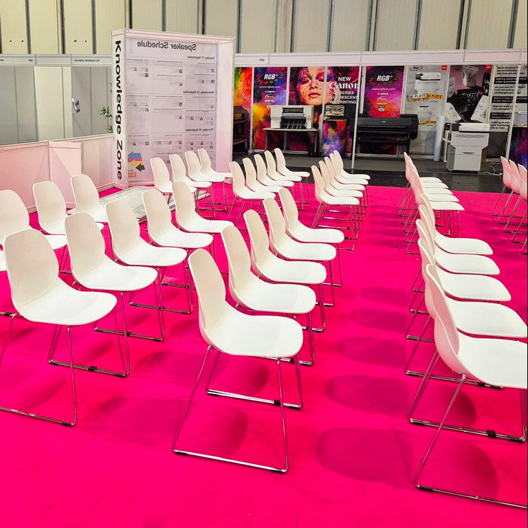 A large exhibition hall floor arranged with rows of white molded plastic chairs facing a stage area with a pink carpet, booths, and display materials.