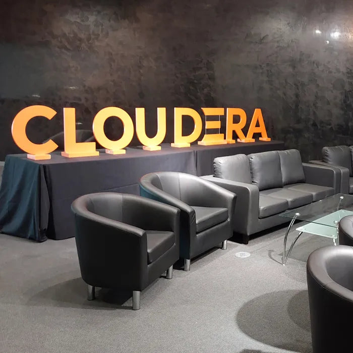 Image of A modern lounge setup with multiple gray leather armchairs and sofas arranged around a small glass coffee table in a dark room. Large illuminated orange letters spelling CLOUDERA are mounted on the wall above a draped table.