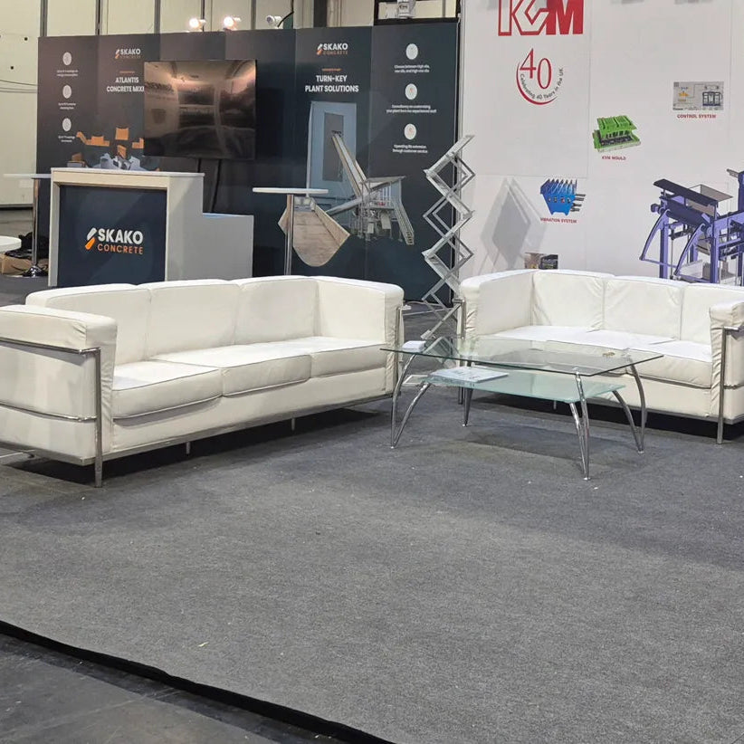 A showroom display featuring two white upholstered sofas with chrome metal frames and a glass-top coffee table on a gray carpet, with industrial machinery and branded booths in the background.