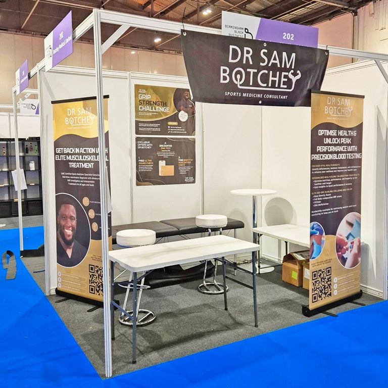 A booth display for 'Dr Sam Botches' sports medicine consultancy at a trade show, featuring white tables and stools, informational banners, and a black hanging sign.