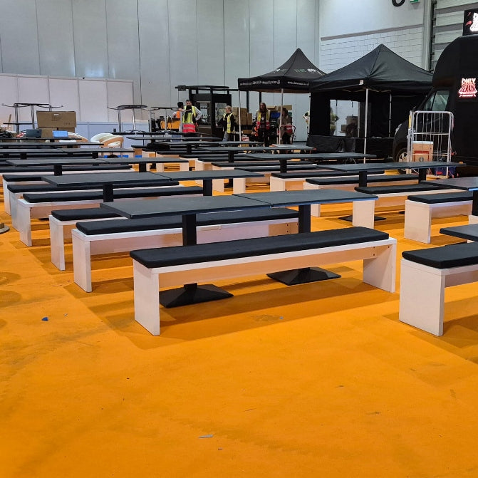 Image of White and black modular outdoor dining tables and benches arranged on a bright orange floor at an event, with a black food truck and tents in the background.