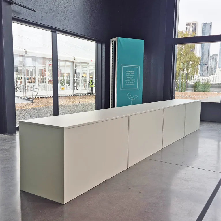 Image of A long, low modern reception desk in a light sage green color, positioned in a contemporary office lobby with large windows showing a city skyline outside.