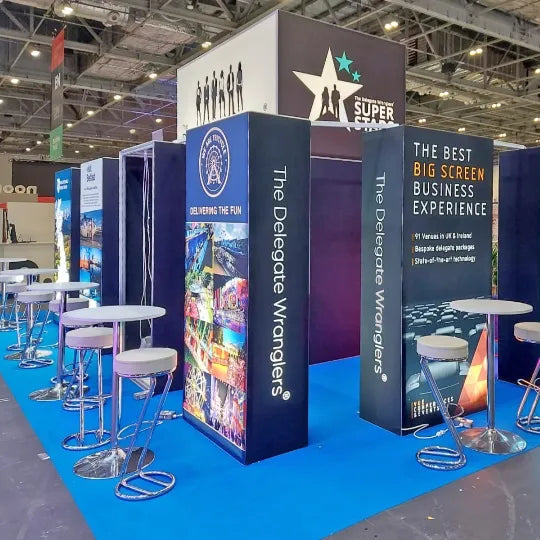 Image of A large exhibition booth setup at a trade show, featuring multiple vertical panels with business-themed graphics and text, round white tables, and metal bar stools on a blue carpeted floor in a convention hall.