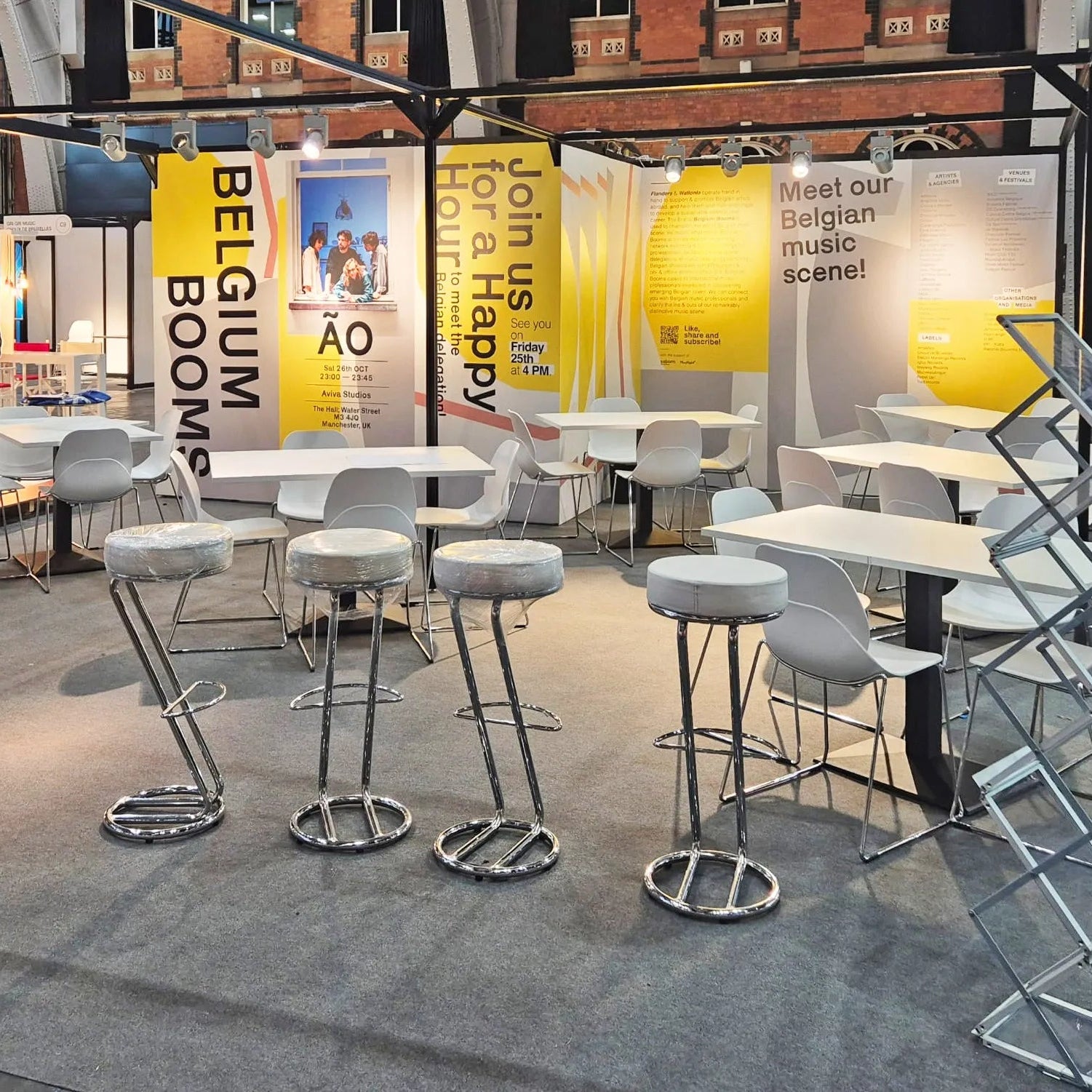 Image of A large exhibition booth displaying modern white bar stools with chrome bases, white tables, and yellow promotional banners reading 'BELGIUM BOOM' and 'Meet our Belgian music scene!'.