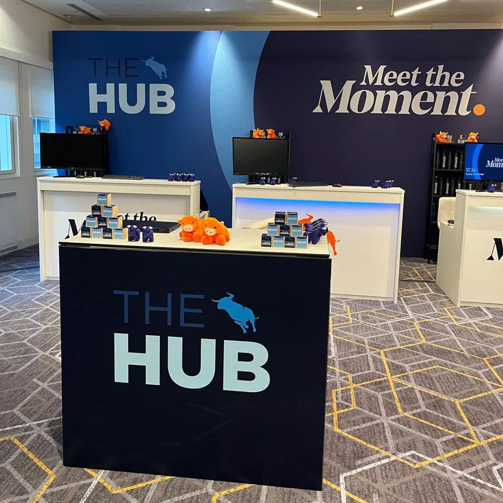 A branded retail display booth for 'THE HUB' featuring a dark navy counter in the foreground, white display tables, computer monitors, and a large blue and white branded wall backdrop in an office-like interior.